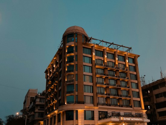 A tall, well-lit hotel building with a modern design stands against a dusky sky. The structure has multiple windows and a prominent signage, suggesting it's a notable establishment. The lighting enhances the warm tones of the building.
