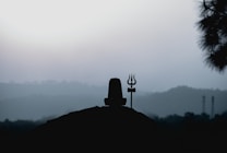 A silhouette of a Shiva Lingam and a trident against a backdrop of misty hills and a pale sky. The scene conveys a sense of tranquility and spirituality.