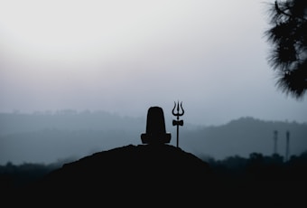 A silhouette of a Shiva Lingam and a trident against a backdrop of misty hills and a pale sky. The scene conveys a sense of tranquility and spirituality.