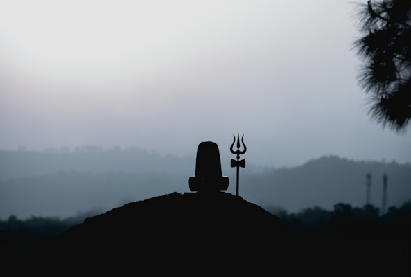 A silhouette of a Shiva Lingam and a trident against a backdrop of misty hills and a pale sky. The scene conveys a sense of tranquility and spirituality.