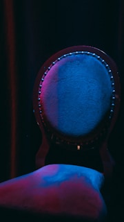 A single, ornately designed chair with a round backrest is illuminated by dramatic lighting. The upholstery appears textured and is bathed in deep, moody hues of blue and purple, casting intriguing shadows that enhance its elegance.
