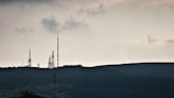 Telecommunication towers are silhouetted against a cloudy sky, standing on a hilly terrain covered with dark vegetation. The scene is calm yet somewhat imposing, with an overcast atmosphere.