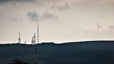 Telecommunication towers are silhouetted against a cloudy sky, standing on a hilly terrain covered with dark vegetation. The scene is calm yet somewhat imposing, with an overcast atmosphere.