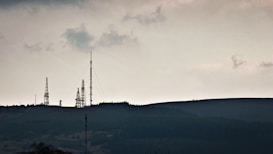 Telecommunication towers are silhouetted against a cloudy sky, standing on a hilly terrain covered with dark vegetation. The scene is calm yet somewhat imposing, with an overcast atmosphere.