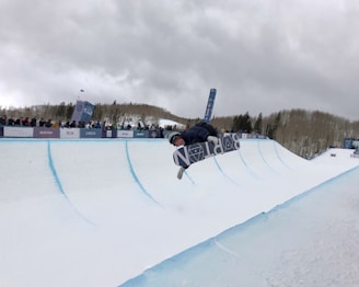 A snowboarder performs a trick mid-air above a snow-covered half-pipe. The snowboarder is wearing a helmet and goggles for protection. A crowd of spectators watches from behind barriers lining the half-pipe, with banners of various sponsors visible. The scene is set in a snowy, forested area under a cloudy gray sky.