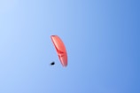 A paraglider is soaring high in the sky, supported by an orange and white parachute. The sky is clear and blue, providing a serene backdrop to the solo glider.
