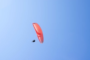 A paraglider is soaring high in the sky, supported by an orange and white parachute. The sky is clear and blue, providing a serene backdrop to the solo glider.