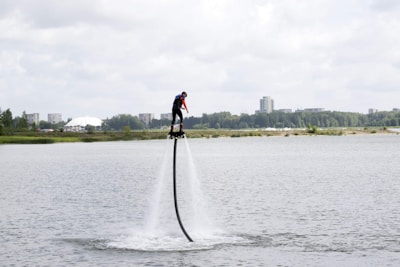 A person is flyboarding over a body of water, propelled by water jets beneath their feet. The background features distant buildings, trees, and a partially cloudy sky.