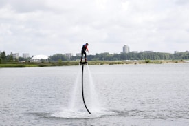 A person is flyboarding over a body of water, propelled by water jets beneath their feet. The background features distant buildings, trees, and a partially cloudy sky.