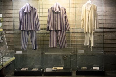 Three sets of clothing are displayed in glass cases against a textured wall. The two garments on the left consist of blue and gray striped fabric, both including a top and pants, suggesting prison uniforms. The garment on the right is a plain, off-white long-sleeved top and matching pants. Various documents or items are placed at the bottom of the cases.