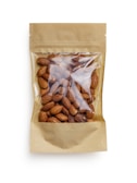 A brown paper bag with a transparent window displaying a collection of whole almonds. The packaging is simple and minimalistic, focusing on the natural appearance of the almonds inside.