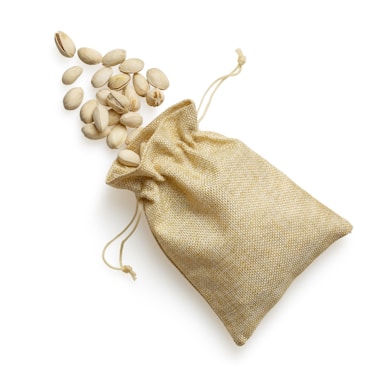 A small burlap sack with a drawstring is placed on a white background. Several pistachio nuts are spilling out of the opening and scattered around.