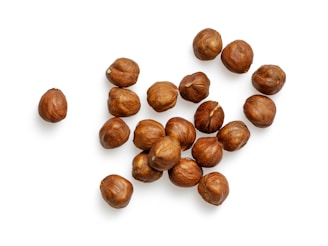 A cluster of hazelnuts scattered on a white background, each nut displaying a rich, brown color with a textured surface.
