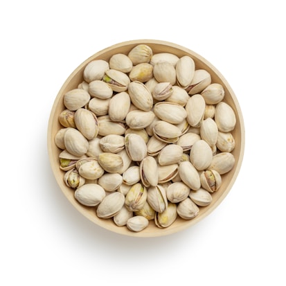 A wooden bowl filled with a pile of pistachio nuts, some of which are slightly open to reveal the greenish nut inside. The bowl is centered against a plain white background.