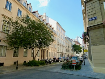 A European street scene features traditional architecture with beige buildings adorned with red-tiled roofs and dormer windows. Lush green trees line the quiet street, where several parked cars and motorcycles can be seen. A signpost indicating a street name is visible on the right, and a clear blue sky overhead completes the setting.
