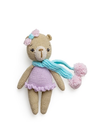 A crocheted teddy bear wearing a pastel purple dress, adorned with a pink bow on its head and a light blue scarf with pom poms. The bear has a beige body and a light brown nose, with dark eyes that give it a cute appearance.