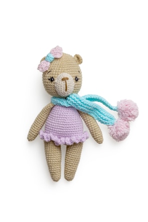 A crocheted teddy bear wearing a pastel purple dress, adorned with a pink bow on its head and a light blue scarf with pom poms. The bear has a beige body and a light brown nose, with dark eyes that give it a cute appearance.