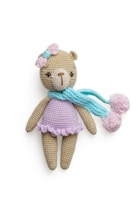 A crocheted teddy bear wearing a pastel purple dress, adorned with a pink bow on its head and a light blue scarf with pom poms. The bear has a beige body and a light brown nose, with dark eyes that give it a cute appearance.