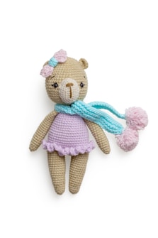A crocheted teddy bear wearing a pastel purple dress, adorned with a pink bow on its head and a light blue scarf with pom poms. The bear has a beige body and a light brown nose, with dark eyes that give it a cute appearance.