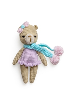 A crocheted teddy bear wearing a pastel purple dress, adorned with a pink bow on its head and a light blue scarf with pom poms. The bear has a beige body and a light brown nose, with dark eyes that give it a cute appearance.