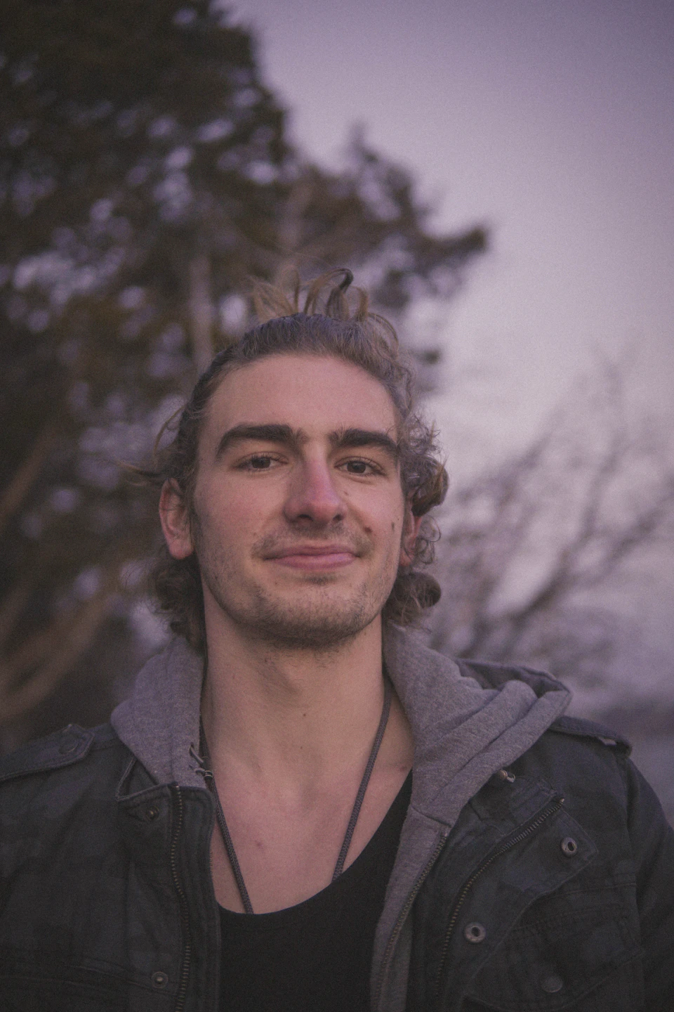 In the image, a young man with curly hair is the main subject. He's wearing a black jacket and has a warm smile on his face. The background features trees and bushes, adding to the natural ambiance of the scene. The photo appears to be taken outdoors during the day under a cloudy sky, giving it an atmospheric quality.