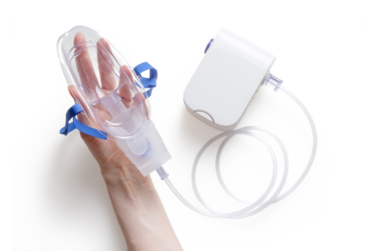a hand holding an oxygen mask that is connected to tubes and a little white machine all against a white background.