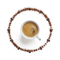 A cup of coffee sits in the center of the image surrounded by a circle made from coffee beans and coffee grounds. The cup is filled with freshly brewed coffee, showing a foamy crema on top. The arrangement of the coffee beans and grounds is symmetrical and gives a feeling of balance and harmony.