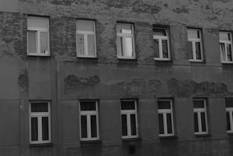 A building facade with visible signs of wear and aging, featuring multiple rows of windows. The wall surface shows peeling paint and exposed patches of brickwork, indicating a lack of maintenance.