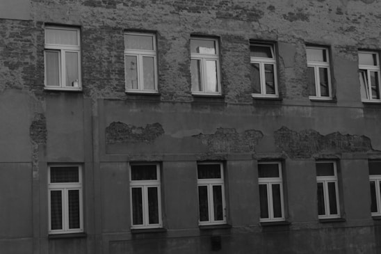 A building facade with visible signs of wear and aging, featuring multiple rows of windows. The wall surface shows peeling paint and exposed patches of brickwork, indicating a lack of maintenance.