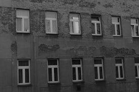 A building facade with visible signs of wear and aging, featuring multiple rows of windows. The wall surface shows peeling paint and exposed patches of brickwork, indicating a lack of maintenance.