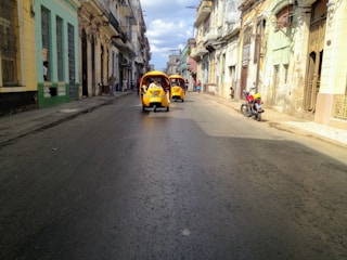 A street scene in a historic or vintage neighborhood with two bright yellow, three-wheeled motorized rickshaws moving down the road. The buildings appear old and slightly rundown, with balconies and various architectural details. A red motorcycle is parked on the sidewalk to the right, and a few pedestrians are seen walking or talking in the distance.