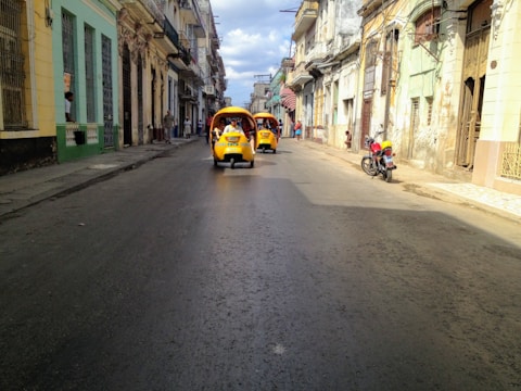 A street scene in a historic or vintage neighborhood with two bright yellow, three-wheeled motorized rickshaws moving down the road. The buildings appear old and slightly rundown, with balconies and various architectural details. A red motorcycle is parked on the sidewalk to the right, and a few pedestrians are seen walking or talking in the distance.