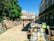 An outdoor book market set up in a sunlit plaza, with multiple stands displaying a variety of books. People are casually browsing the selections, enjoying the warm, inviting atmosphere. Surrounding the market are elegant, historical buildings featuring arched windows and balconies. A large tree provides shade on part of the cobblestone ground.