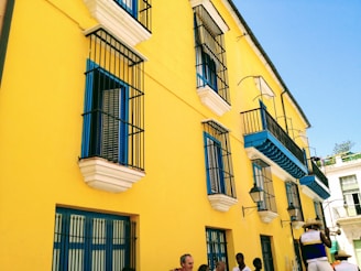 A vibrant yellow building with blue and white accents features multiple windows with black wrought iron bars. Several people are standing on the sidewalk, and a musician is playing a trumpet, wearing a hat and colorful clothing. The sky is clear and blue.