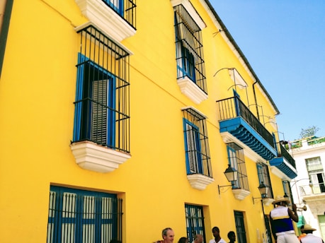 A vibrant yellow building with blue and white accents features multiple windows with black wrought iron bars. Several people are standing on the sidewalk, and a musician is playing a trumpet, wearing a hat and colorful clothing. The sky is clear and blue.