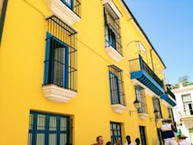 A vibrant yellow building with blue and white accents features multiple windows with black wrought iron bars. Several people are standing on the sidewalk, and a musician is playing a trumpet, wearing a hat and colorful clothing. The sky is clear and blue.