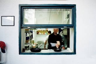 A chef is working in a kitchen, visible through a window with a blue frame. Several pans are on the counter. The walls are white, and there are various kitchen supplies and ingredients visible on shelves in the background.