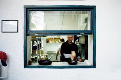 A chef is working in a kitchen, visible through a window with a blue frame. Several pans are on the counter. The walls are white, and there are various kitchen supplies and ingredients visible on shelves in the background.