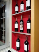 A red shelf holds eight bottles of wine, each with a label. The arrangement is neat with three rows and varying bottle placements. A window is visible to the left, allowing natural light.