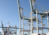 Massive industrial cranes dominate a port scene, with complex metal structures and machinery visible against a clear blue sky. These cranes are positioned above shipping containers and are likely used for loading and unloading cargo from ships.