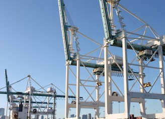 Massive industrial cranes dominate a port scene, with complex metal structures and machinery visible against a clear blue sky. These cranes are positioned above shipping containers and are likely used for loading and unloading cargo from ships.