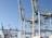 Massive industrial cranes dominate a port scene, with complex metal structures and machinery visible against a clear blue sky. These cranes are positioned above shipping containers and are likely used for loading and unloading cargo from ships.