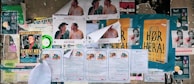 A collage of various posters and flyers are affixed to a bulletin board. The posters feature different advertisements, primarily in bold and colorful fonts. Some depict illustrations of individuals or items, while others include text-heavy information. The edges of certain papers are curling or partially torn, indicating exposure to weather or frequent re-pasting. The visual clutter creates a busy, layered look.