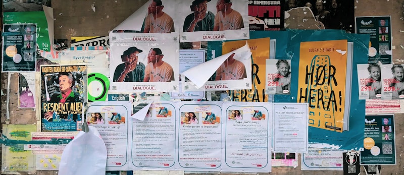 A collage of various posters and flyers are affixed to a bulletin board. The posters feature different advertisements, primarily in bold and colorful fonts. Some depict illustrations of individuals or items, while others include text-heavy information. The edges of certain papers are curling or partially torn, indicating exposure to weather or frequent re-pasting. The visual clutter creates a busy, layered look.