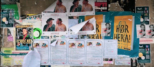 A collage of various posters and flyers are affixed to a bulletin board. The posters feature different advertisements, primarily in bold and colorful fonts. Some depict illustrations of individuals or items, while others include text-heavy information. The edges of certain papers are curling or partially torn, indicating exposure to weather or frequent re-pasting. The visual clutter creates a busy, layered look.