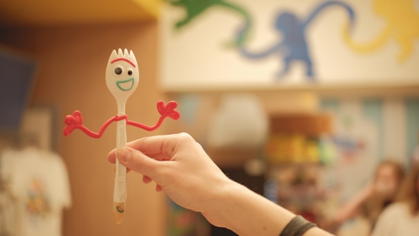 A hand holds a toy resembling a spork with googly eyes and red pipe cleaner arms. The toy has a cheerful face with a wide smile, and the background appears to be a store with colorful wall art depicting monkeys.