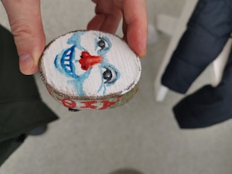 A hand is holding a small round piece of wood with a painted clown face on one side. The clown has a blue and white face with a red nose and a blue smile, along with black detailed eyes. In the background, there is part of a black jacket and textured flooring.