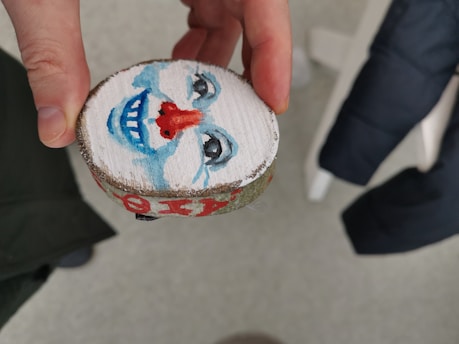 A hand is holding a small round piece of wood with a painted clown face on one side. The clown has a blue and white face with a red nose and a blue smile, along with black detailed eyes. In the background, there is part of a black jacket and textured flooring.