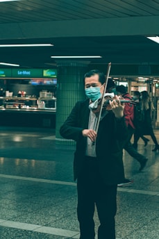 A person wearing a face mask is playing a violin in an indoor public space with tiled flooring. The background shows a food counter and several people walking by.