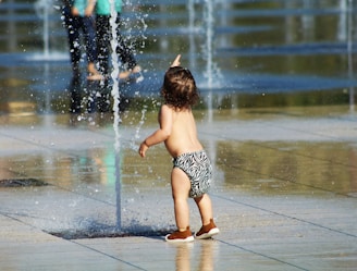 A young child in a patterned diaper is playing in an area with water fountains, pointing towards the water jets. The surroundings are wet, reflecting the sunlight creating a playful and lively atmosphere.